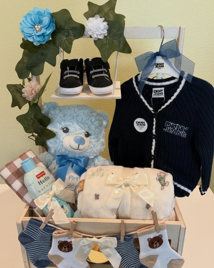 Collection of baby items including a teddy bear, shoes, and clothing in a decorative box with flowers.